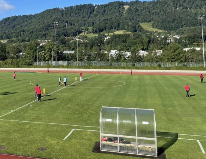 A wide green ground with a wooded hillside behind it, players spread in the field